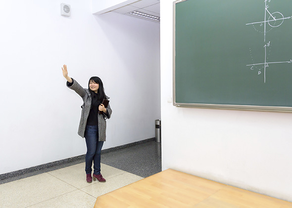 Asian teacher waves near a green chalkboard