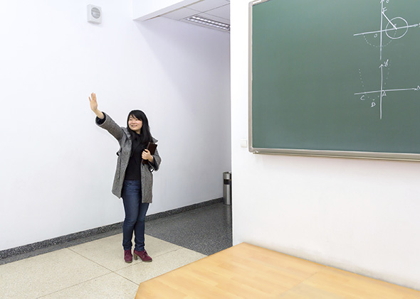 150304_HIED_TeachBias Asian teacher waves near a green chalkboard