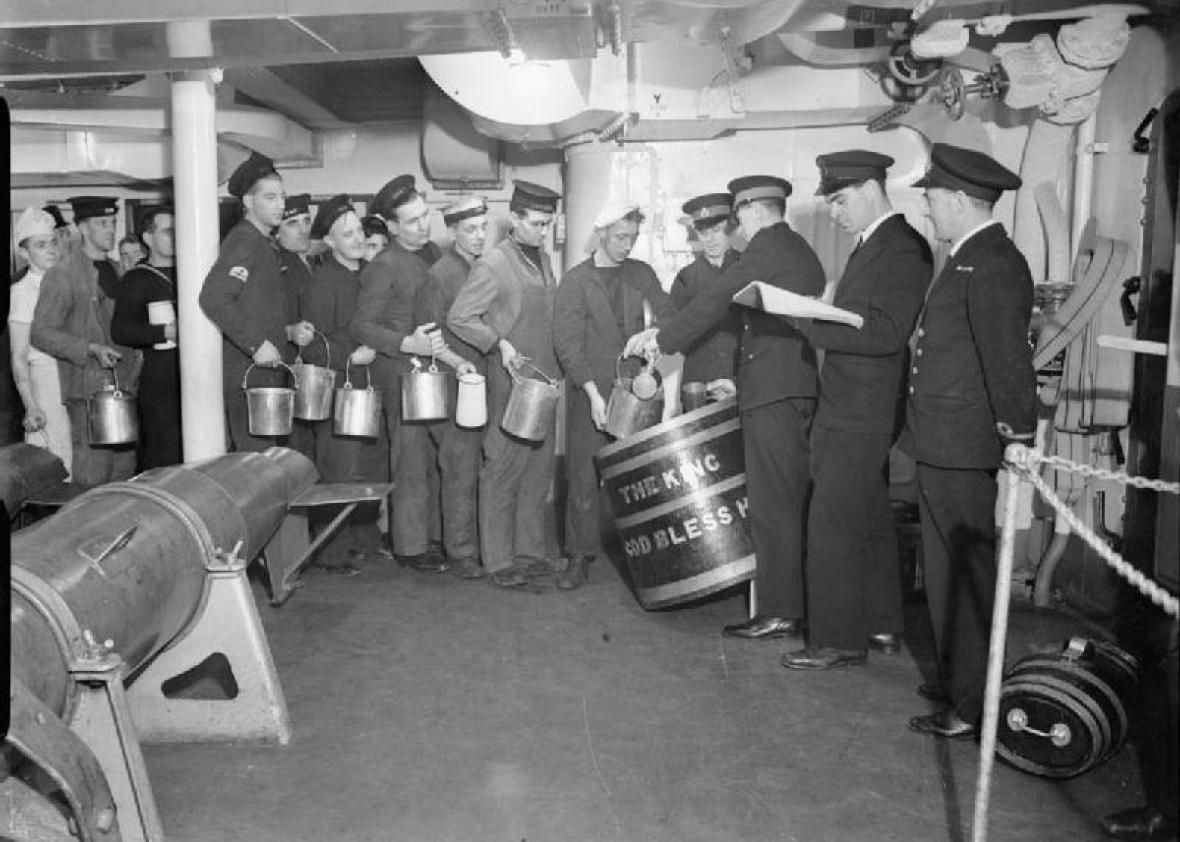 Below deck, a line of seamen queue to collect the daily rum ration for their mess. Each man is holding a jug or bucket. The rum is being issued from a large barrel with 'THE KING - GOD BLESS HIM' on it. Royal Marines issue the rum with measuring jugs while a Royal Navy Petty Officer and Sub-Lieutenant observe.