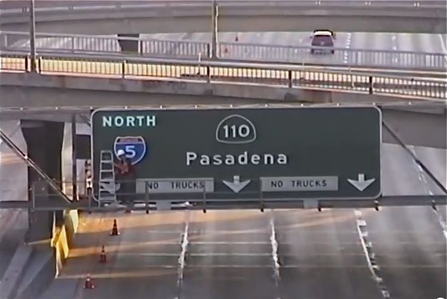 A man in a construction hat and vest hangs an "Interstate 5" shield on a freeway signboard.