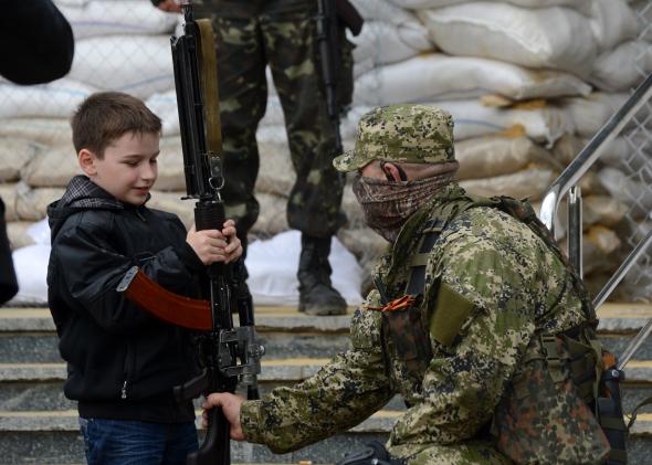 485746183-young-boy-holds-the-machine-gun-of-an-armed-man-in