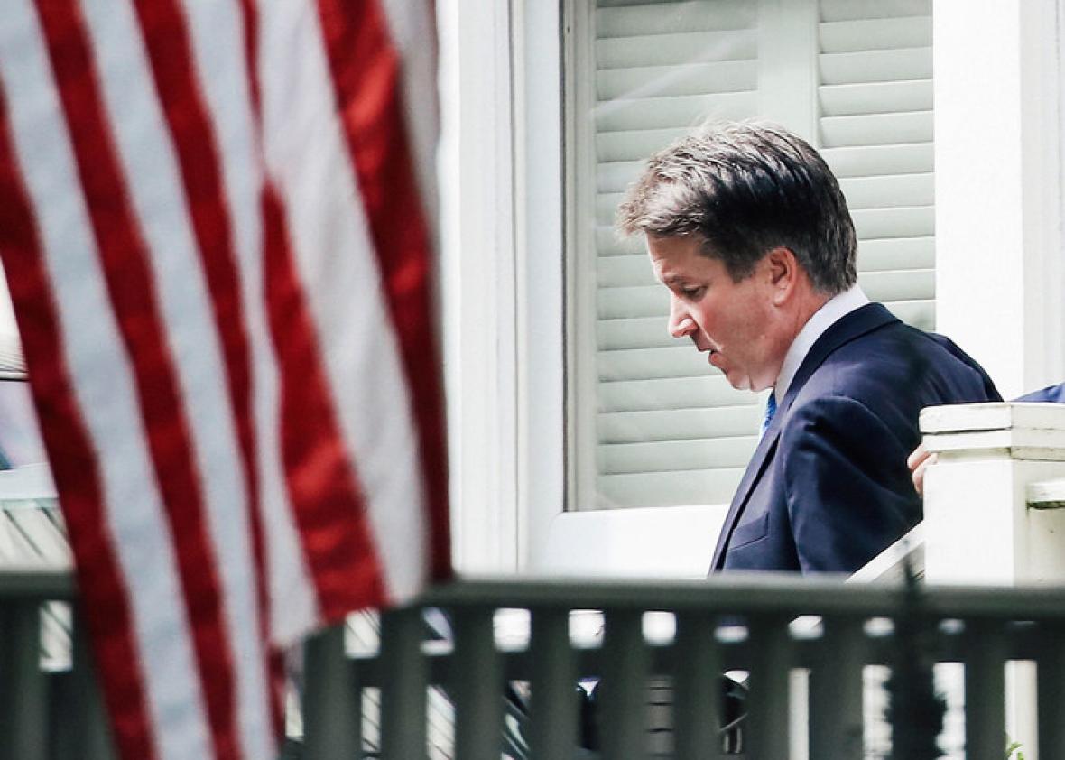Brett Kavanaugh, eyes downcast, walking out of his home, with an American flag in the foreground.